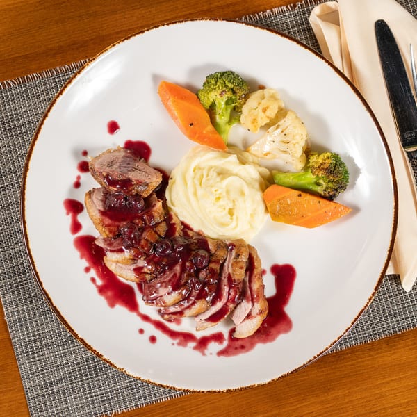 Sliced duck breast with berry sauce, mashed potatoes, and roasted vegetables on a white plate.