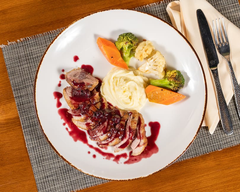 Sliced duck breast with berry sauce, mashed potatoes, and roasted vegetables on a white plate.