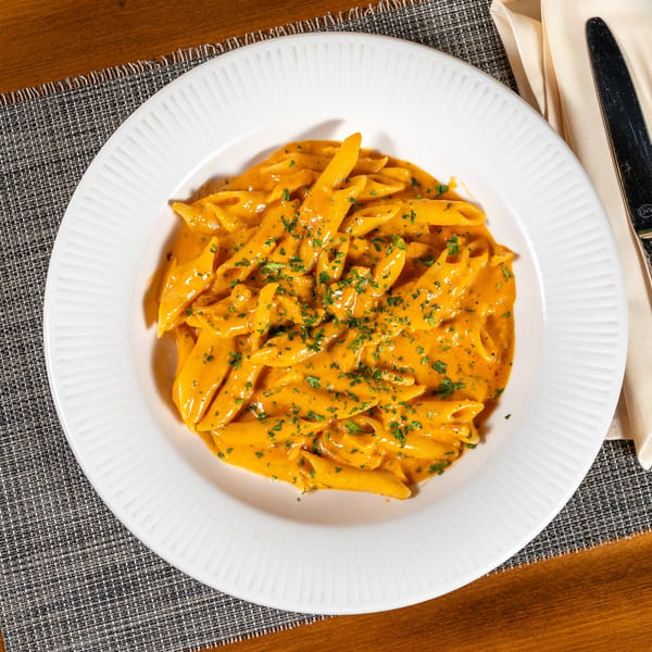 Penne pasta in a creamy orange sauce, garnished with parsley, on a white plate with cutlery.