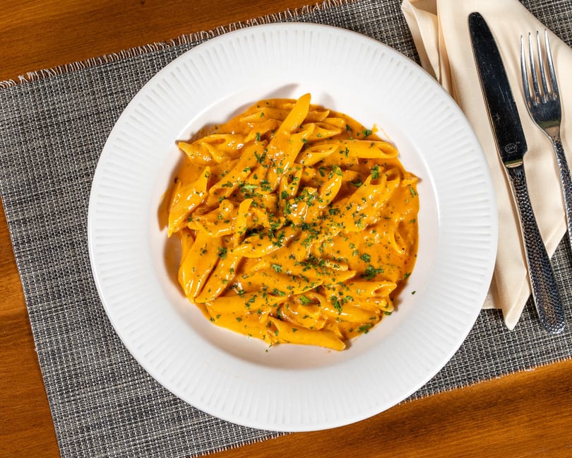 Penne pasta in a creamy orange sauce, garnished with parsley, on a white plate with cutlery.