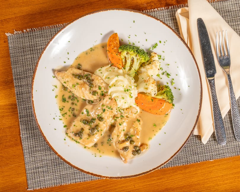 Chicken piccata with capers, mashed potatoes, broccoli, and carrots on a white plate.