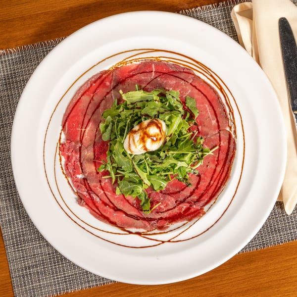 Carpaccio appetizer with thinly sliced meat, arugula, burrata cheese, and balsamic glaze on a white plate.