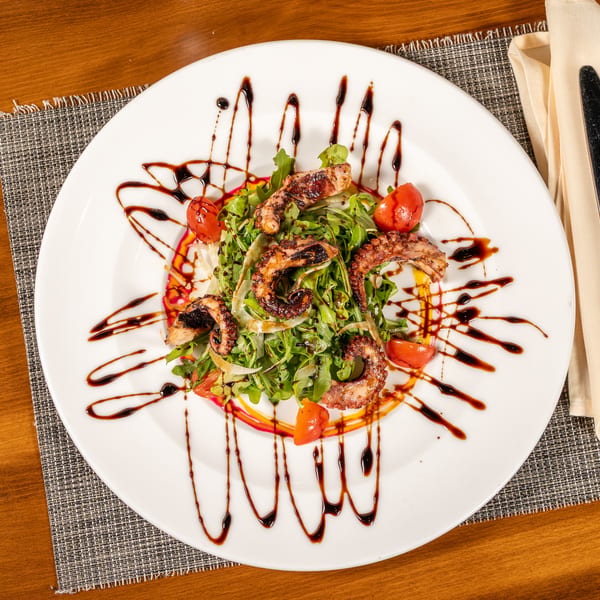 Grilled octopus salad with arugula, cherry tomatoes, and balsamic glaze on a white plate.