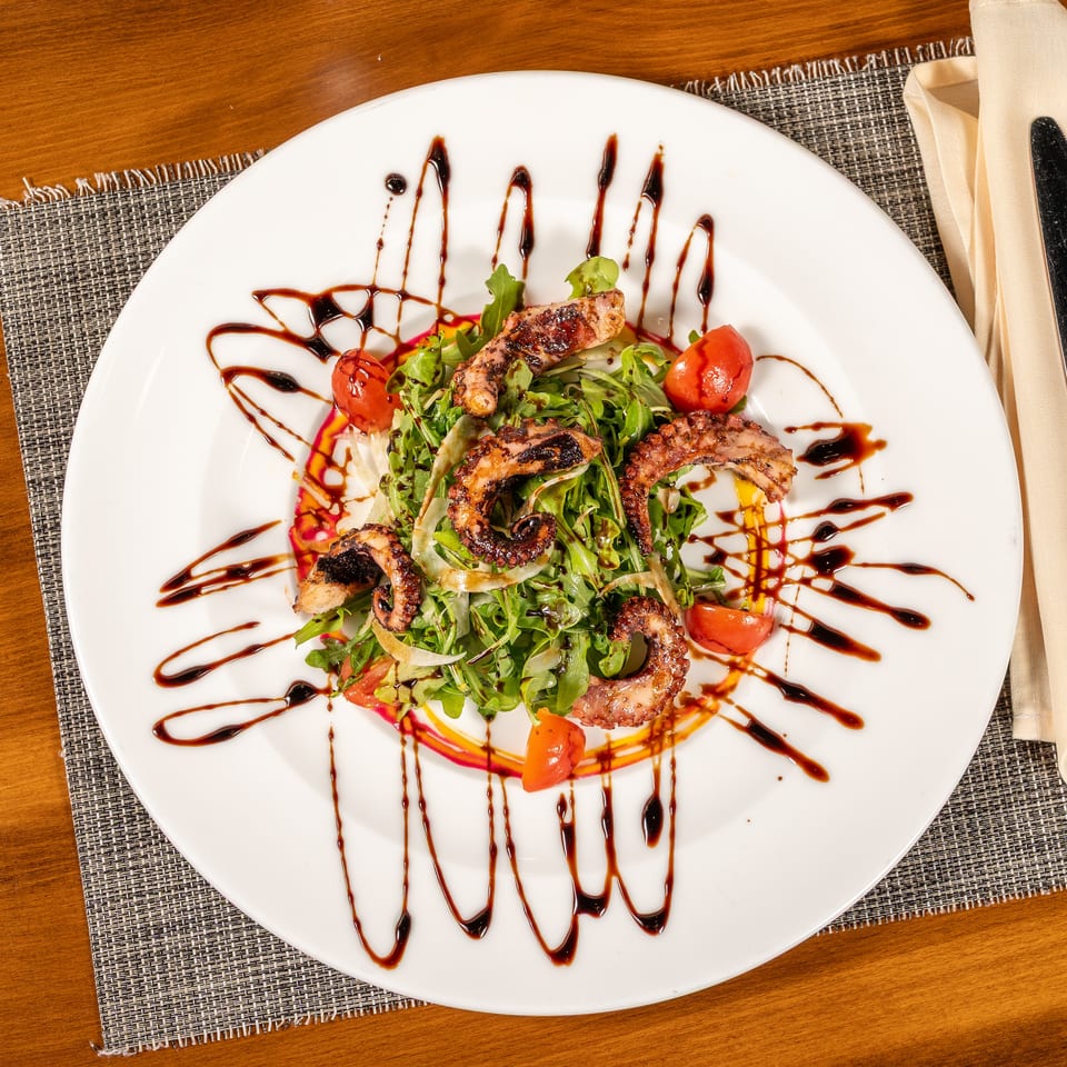 Grilled octopus salad with arugula, cherry tomatoes, and balsamic glaze on a white plate.