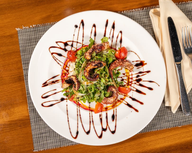 Grilled octopus salad with arugula, cherry tomatoes, and balsamic glaze on a white plate.