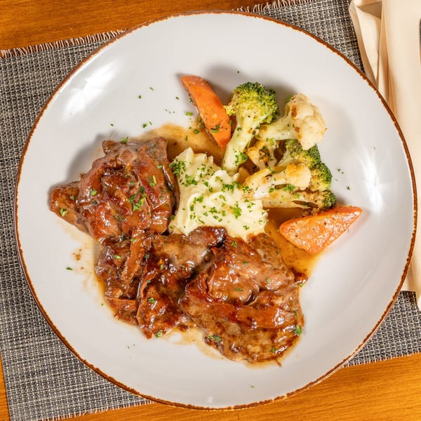 Savory meat dish with gravy, mashed potatoes, broccoli, cauliflower, and carrots on a white plate.