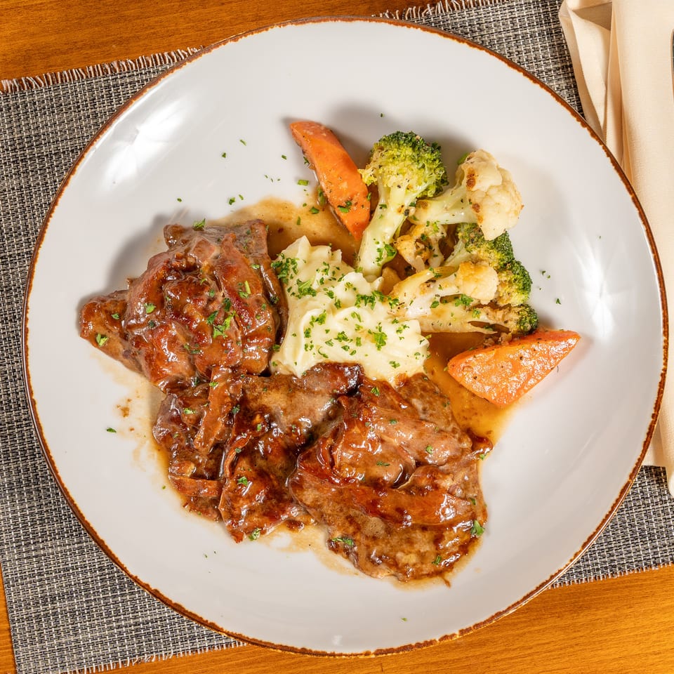 Savory meat dish with gravy, mashed potatoes, broccoli, cauliflower, and carrots on a white plate.