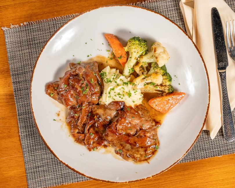 Savory meat dish with gravy, mashed potatoes, broccoli, cauliflower, and carrots on a white plate.