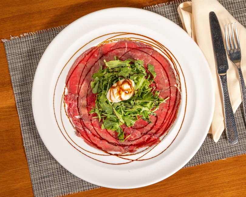 Carpaccio with arugula, burrata cheese, and balsamic glaze on a white plate with cutlery.